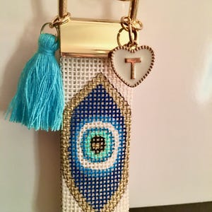 May include: A white and gold key chain with a blue evil eye charm, a turquoise tassel, and a gold heart charm with the letter "T" on it.