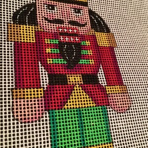 May include: A colorful needlepoint design of a nutcracker soldier. The figure is rendered in a pixelated style, with a red jacket, green pants, and a black hat. The design is on a white grid background.