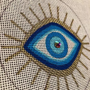 May include: A needlepoint canvas featuring an eye design stitched in concentric circles of dark blue, medium blue, and light blue thread. The eye is surrounded by radiating gold metallic rays and features a central iridescent crystal embellishment.