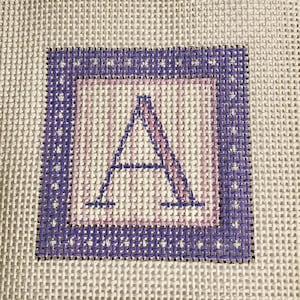 May include: Needlepoint canvas featuring a stitched letter "A" in a square design. The letter is in a light pink color, framed by a light pink square, and a dark purple border. The background is a white canvas.