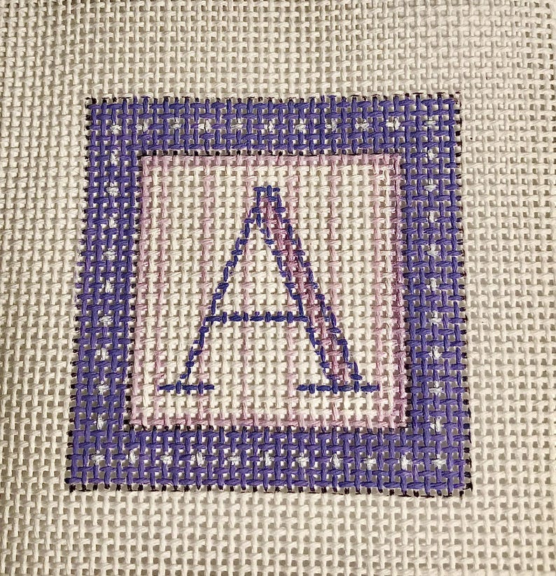 May include: Needlepoint canvas featuring a stitched letter "A" in a square design. The letter is in a light pink color, framed by a light pink square, and a dark purple border. The background is a white canvas.