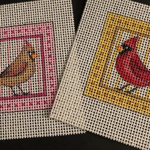 May include: Two needlepoint canvases featuring cardinal birds. One canvas has a brown bird in a pink frame, the other has a red bird in a yellow frame. Both are on a white mesh background.