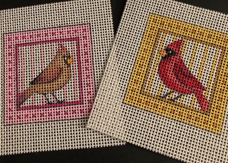 May include: Two needlepoint canvases featuring cardinal birds. One canvas has a brown bird in a pink frame, the other has a red bird in a yellow frame. Both are on a white mesh background.
