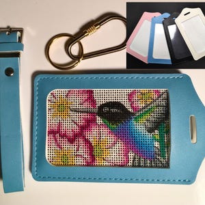 May include: A light blue luggage tag with a stitched hummingbird design, a matching strap, and a gold-colored carabiner clip. Additional luggage tags in pink, blue, black, and white are also shown.