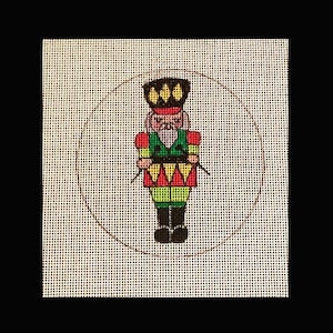 May include: A round canvas with a brown outline, featuring a colorful nutcracker holding a drum. The nutcracker is wearing a red and green uniform with a black hat and yellow accents.