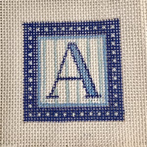 May include: Needlepoint canvas featuring the letter "A" in shades of blue. The letter is centered within a light blue and dark blue square border, with a white background. The canvas is made of a woven material.
