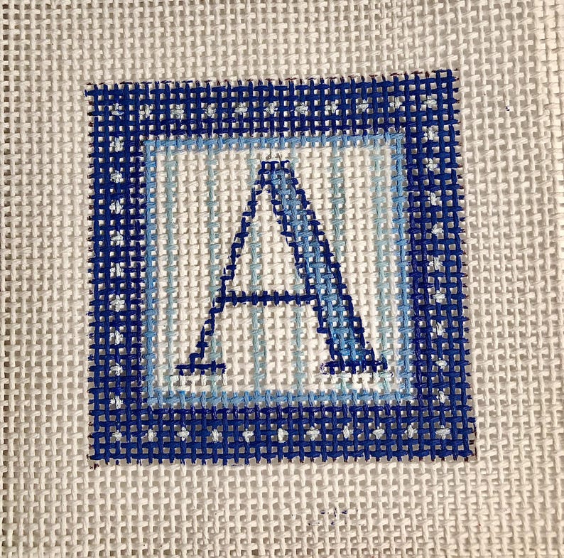 May include: Needlepoint canvas featuring the letter "A" in shades of blue. The letter is centered within a light blue and dark blue square border, with a white background. The canvas is made of a woven material.