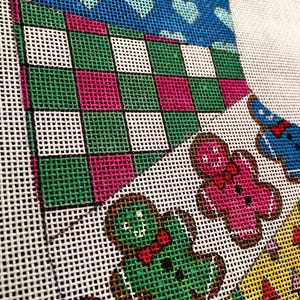 May include: A close-up of a needlepoint canvas with a colorful design. The design features a green, pink, and white checkered pattern, a blue section with white hearts, and a green gingerbread man with a red bow tie.