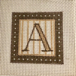 May include: Square needlepoint canvas featuring the letter "A" in a brown, tan, and white color scheme. The letter is centered within a tan and brown border, with a brown outer border. The background is a neutral, off-white color.