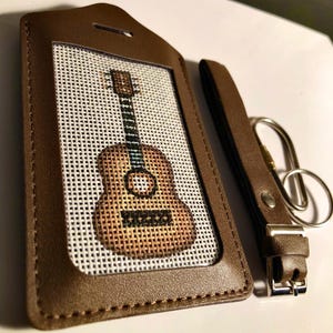 May include: A brown leather luggage tag with a stitched guitar design on a white background. The tag includes a matching strap with a metal clasp and key ring. The guitar is a light brown color.