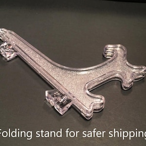 May include: A clear, plastic folding stand with a textured surface. The stand is designed to hold items securely. The text "Folding stand for safer shipping" is visible at the bottom of the image.