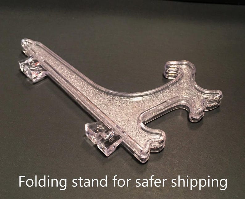 May include: A clear, plastic folding stand with a textured surface. The stand is designed to hold items securely. The text "Folding stand for safer shipping" is visible at the bottom of the image.