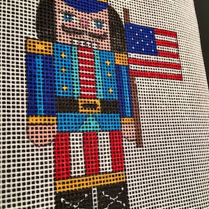 May include: A colorful, pixelated needlepoint design of a nutcracker soldier holding a small American flag. The soldier wears a blue uniform with gold accents, red and white striped pants, and black boots. The background is a white grid.