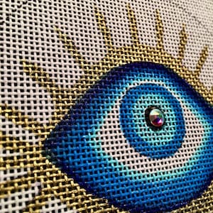 May include: A close-up of a blue and gold evil eye design on a white canvas. The eye is surrounded by gold rays and has a blue and white swirl pattern. A small, clear rhinestone is in the center of the eye.