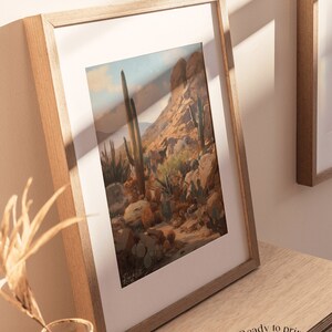 May include: A framed print of a desert landscape with tall cacti and a rocky mountain in the background. The print is in a light wood frame and is hanging on a wall.