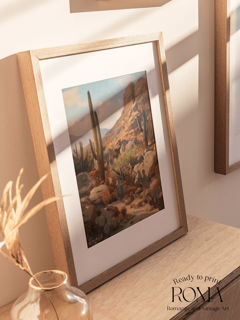 May include: A framed print of a desert landscape with tall cacti and a rocky mountain in the background. The print is in a light wood frame and is hanging on a wall.