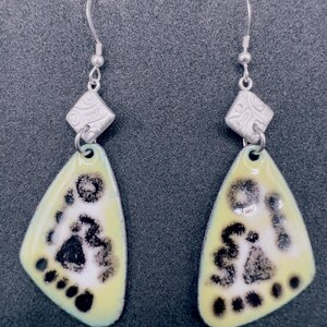 May include: A pair of dangle earrings featuring silver-toned hooks and a textured diamond-shaped silver bead above a triangular pale yellow enamel pendant. The pendant displays a black speckled pattern resembling a footprint with five distinct toe marks.