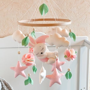Pode incluir: Um móbile para bebê rosa e branco com um coelho dormindo, nuvens, estrelas e flores. O móbile está pendurado em um aro de madeira.