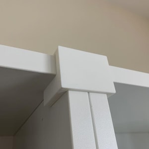 Può includere: Primo piano di un angolo bianco ad angolo retto di un armadio o di una mensola. L'angolo è costruito con linee pulite e un design minimalista. Lo sfondo è di colore beige neutro.