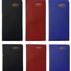 May include: A collection of six rectangular pocket diaries in black, red, and blue. Each diary features a smooth cover, gold-toned metal corner protectors, and the year 2027 embossed in a gold oval frame at the top centre of the front cover.