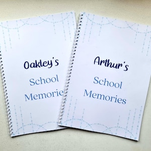 May include: Two white spiral bound notebooks with blue and white patterned covers. The covers have the text "Oakley's School Memories" and "Arthur's School Memories".