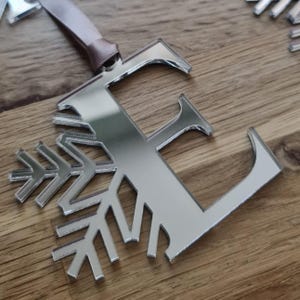 May include: A silver mirrored Christmas decoration combining the letter 'E' with a snowflake design. It hangs from a light brown ribbon. The background is a wooden surface. The ornament is approximately 10 cm in length.