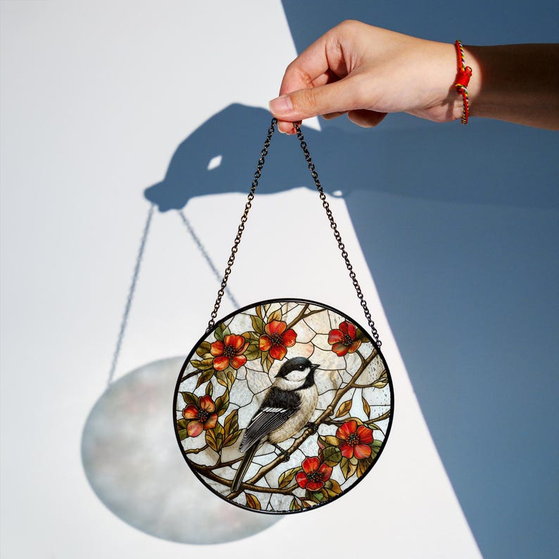 Puede incluir: Un atrapasol de vidrio tintado con un p&aacute;jaro blanco y negro posado en una rama con flores rojas. El atrapasol es redondo y cuelga de una cadena.