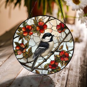 Puede incluir: Una ventana de vidrio emplomado colgante presenta un p&aacute;jaro blanco y negro posado en una rama con flores rojas. El fondo es un vidrio iridiscente blanco y dorado.