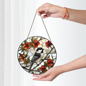 Puede incluir: Una ventana de vidrio emplomado colgada de una cadena. La ventana presenta un p&aacute;jaro blanco y negro posado en una rama con flores rojas. El fondo es un mosaico de blanco, marr&oacute;n y verde.