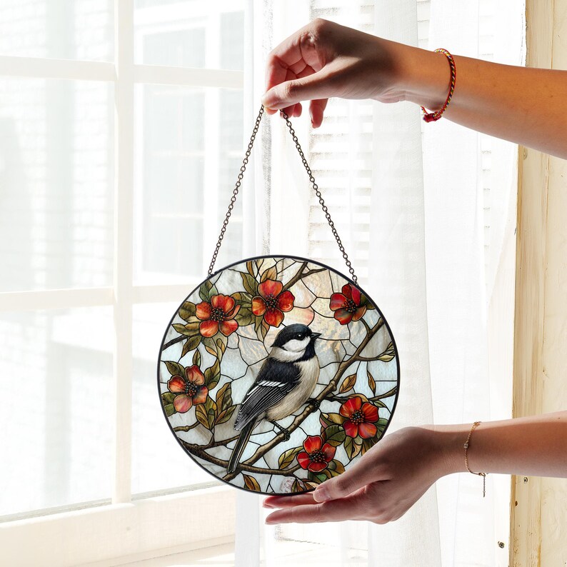 Puede incluir: Una ventana redonda de vidrio emplomado colgada de una cadena. La ventana presenta un p&aacute;jaro blanco y negro posado en una rama con flores rojas. El fondo es de color azul claro con un patr&oacute;n de cuadr&iacute;cula blanca.
