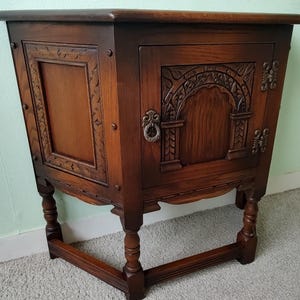 May include: A dark brown wooden side table featuring a single cabinet door with an arched carved panel, a metal ring pull handle, and decorative metal hinges. The table has four turned legs connected by a lower stretcher and carved side panels.