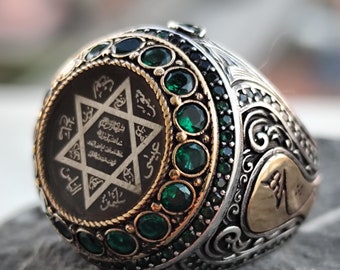 May include: A silver ring with a black and gold star of David design surrounded by green gemstones. The ring has intricate details and a gold band.