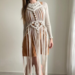 Könnte beinhalten: Eine weiße Makramee-Weste mit langen Fransen, getragen über einem braunen Cordkleid. Die Weste hat ein geometrisches Muster und ist aus dickem Baumwollkordel gefertigt.