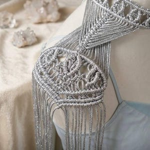 Könnte beinhalten: Ein silbernes Makramee-Schulterstück mit aufwendigen Webmustern und langen Fransen. Das handgefertigte Accessoire wird auf einer Schaufensterpuppe präsentiert, die sein zartes Design und seine Textur hervorhebt. Ein einzigartiges Modeaccessoire.