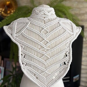 May include: An off-white macrame collar featuring intricate knotted patterns in a chevron design. The piece has a high neck and a pointed hem, constructed from thick cotton cord with a structured, symmetrical arrangement of diagonal and vertical knots.