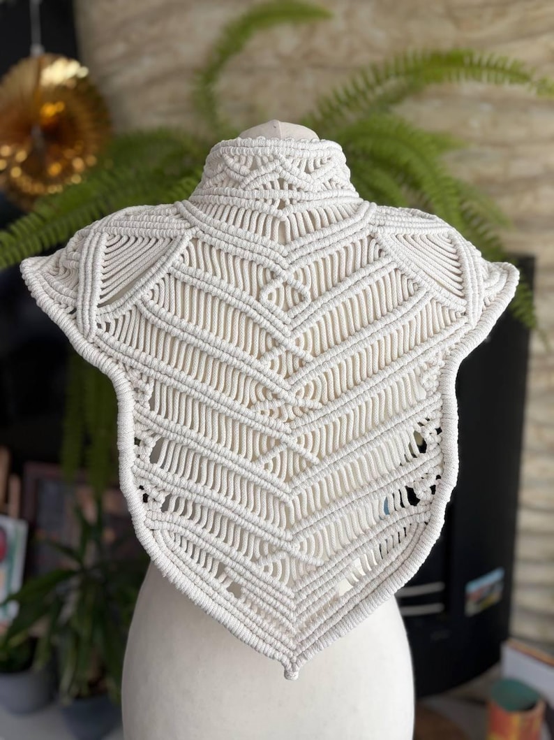 May include: An off-white macrame collar featuring intricate knotted patterns in a chevron design. The piece has a high neck and a pointed hem, constructed from thick cotton cord with a structured, symmetrical arrangement of diagonal and vertical knots.