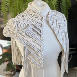 May include: A white crocheted shawl with a geometric pattern and fringe. The shawl is draped over a white mannequin.