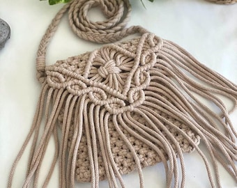 May include: A beige macrame crossbody bag with a long fringe. The bag has a knotted design and a long strap.