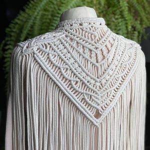 Può includere: Un top bianco all'uncinetto con un design a frangia. Il top ha un'alta scollatura e uno schema geometrico.