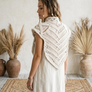 May include: An off-white macram&eacute; vest featuring an intricate chevron pattern and a high collar, worn over a simple, sleeveless, light-colored dress. The vest is crafted from thick, knotted cord and covers the upper back and shoulders.