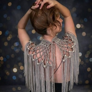 Peut inclure: Écharpe d'épaule argentée à franges, avec un col montant et des détails en macramé. L'écharpe est ornée de petites perles blanches et de longues franges fluides. Le vêtement est porté sur un haut noir, créant un contraste saisissant.