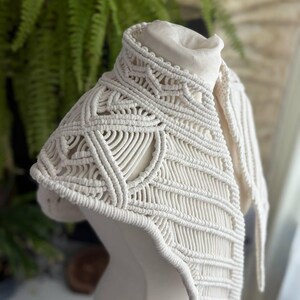 May include: An off-white macrame collar featuring intricate knotted patterns and geometric designs. The accessory is constructed from thick cotton cord, showcasing a high neck structure with detailed woven textures that drape over the shoulders.