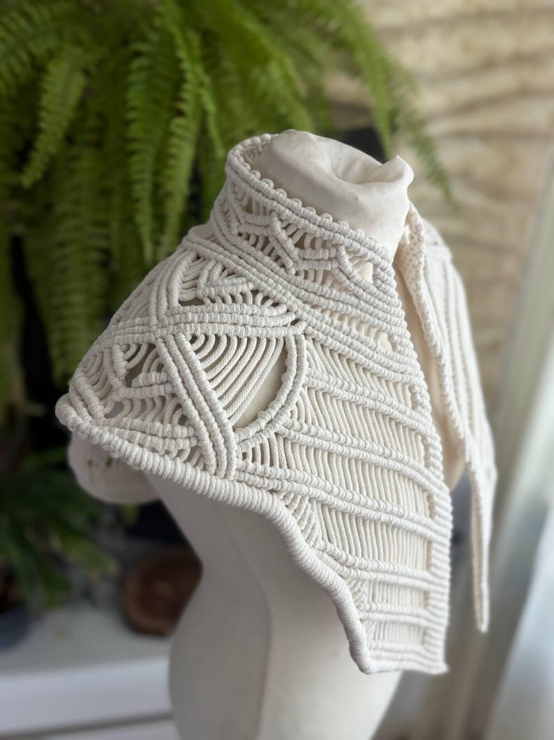 May include: An off-white macrame collar featuring intricate knotted patterns and geometric designs. The accessory is constructed from thick cotton cord, showcasing a high neck structure with detailed woven textures that drape over the shoulders.