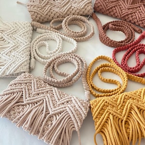Op de afbeelding: Vijf gehaakte tassen met franjes in verschillende kleuren, waaronder wit, beige, bruin, geel en rood. Elke tas heeft een bijpassende gehaakte band.
