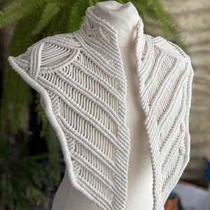 May include: An off-white macrame collar featuring intricate knotted patterns and a high neck design. The piece is constructed from thick cord, showcasing geometric diagonal lines and structured edges that drape over the shoulders and chest area.