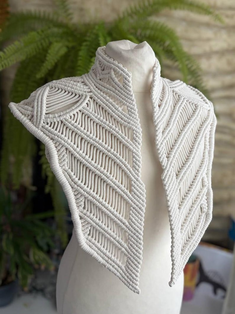 May include: An off-white macrame collar featuring intricate knotted patterns and a high neck design. The piece is constructed from thick cord, showcasing geometric diagonal lines and structured edges that drape over the shoulders and chest area.