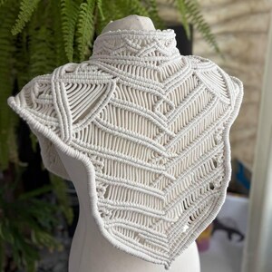 May include: An off-white macrame collar featuring an intricate, hand-knotted geometric pattern. The piece has a high neck design with structured, layered cord work that extends over the shoulders and chest in a pointed, symmetrical shape.
