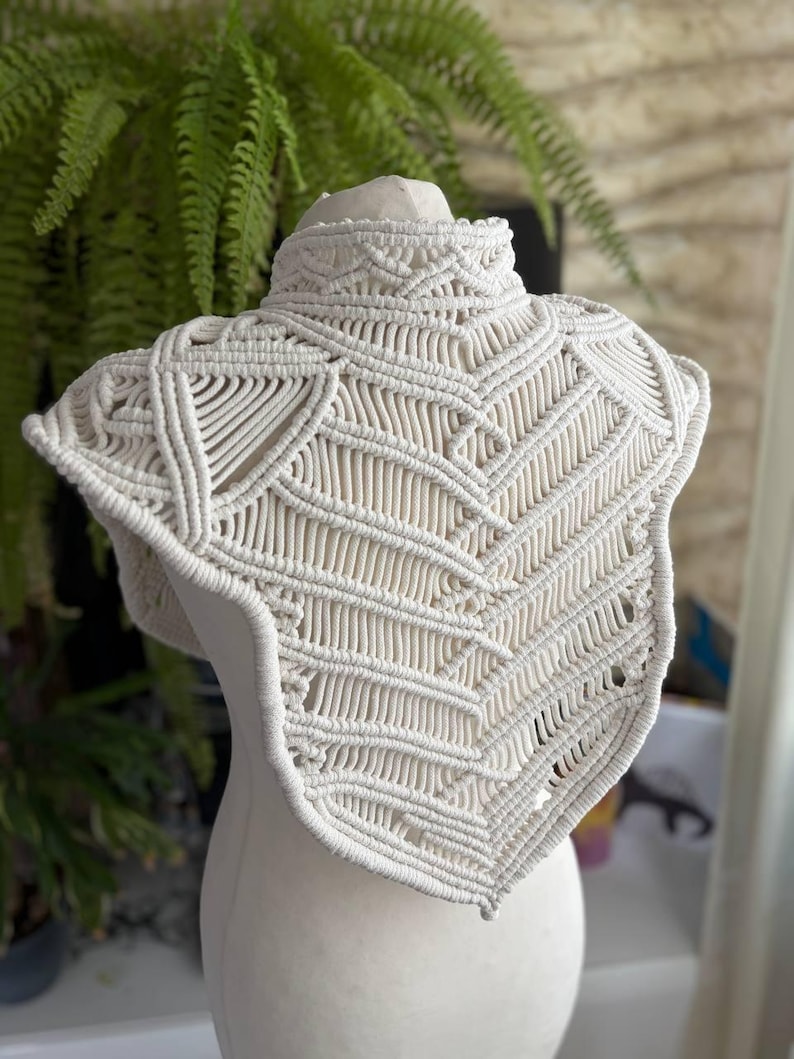 May include: An off-white macrame collar featuring an intricate, hand-knotted geometric pattern. The piece has a high neck design with structured, layered cord work that extends over the shoulders and chest in a pointed, symmetrical shape.