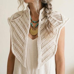 May include: An off-white macrame collar featuring intricate geometric knot patterns and a structured high neckline. The piece is layered over a plain light-colored garment and is paired with a turquoise beaded necklace and a large gold pendant.