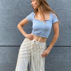May include: A woman wearing a light blue cropped top with a sweetheart neckline and a white crocheted skirt with long fringe.